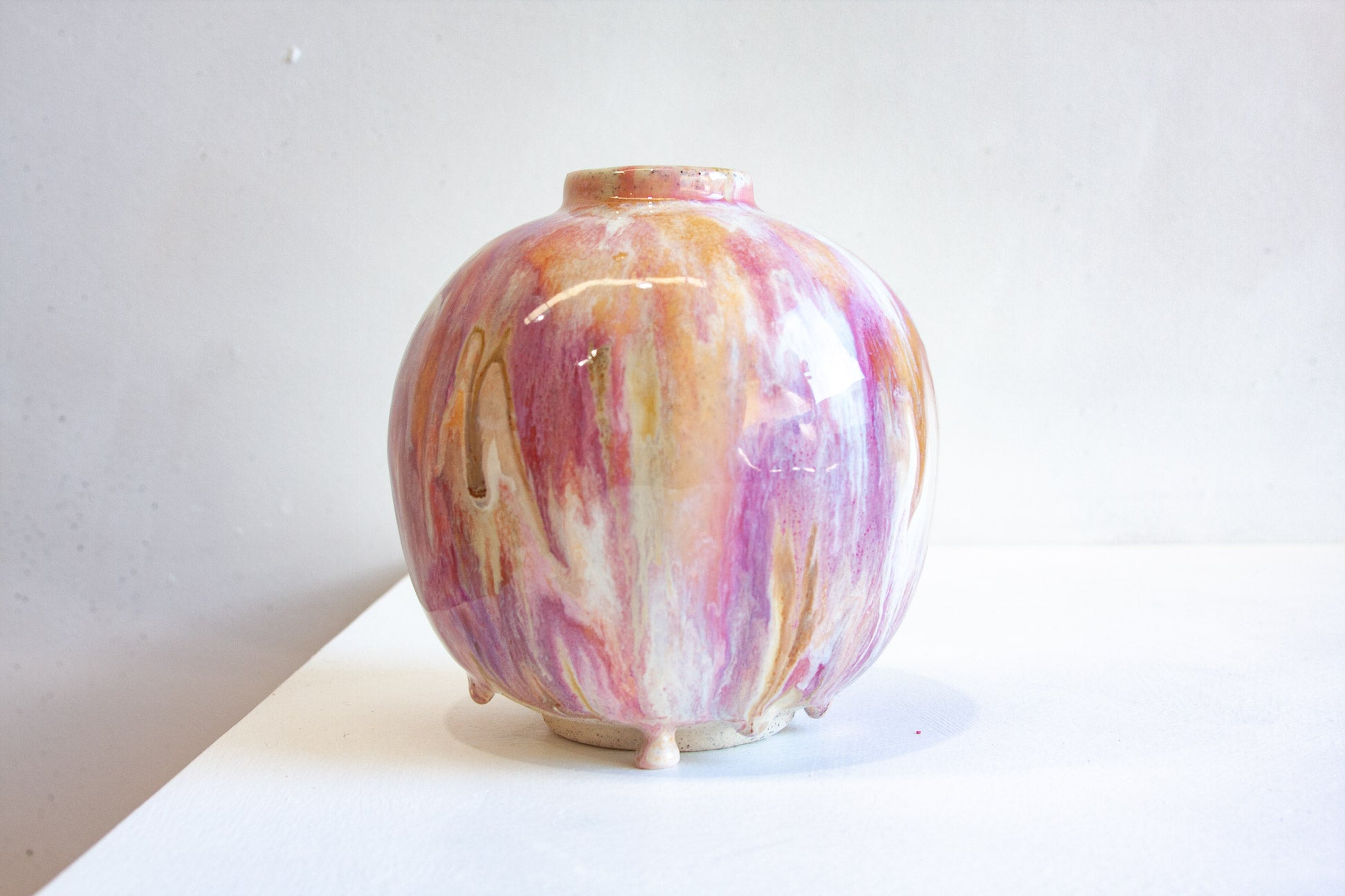 A small round ceramic vase with a short neck and three tiny feet, decorated with a marbled glaze in pink, purple, orange, and cream, set against a plain light gray background.