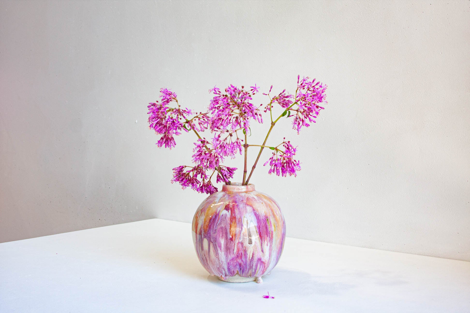 A small round ceramic vase with a marbled pink, purple, and white pattern, holding pink-purple flowers, set on a white surface against a light gray background.