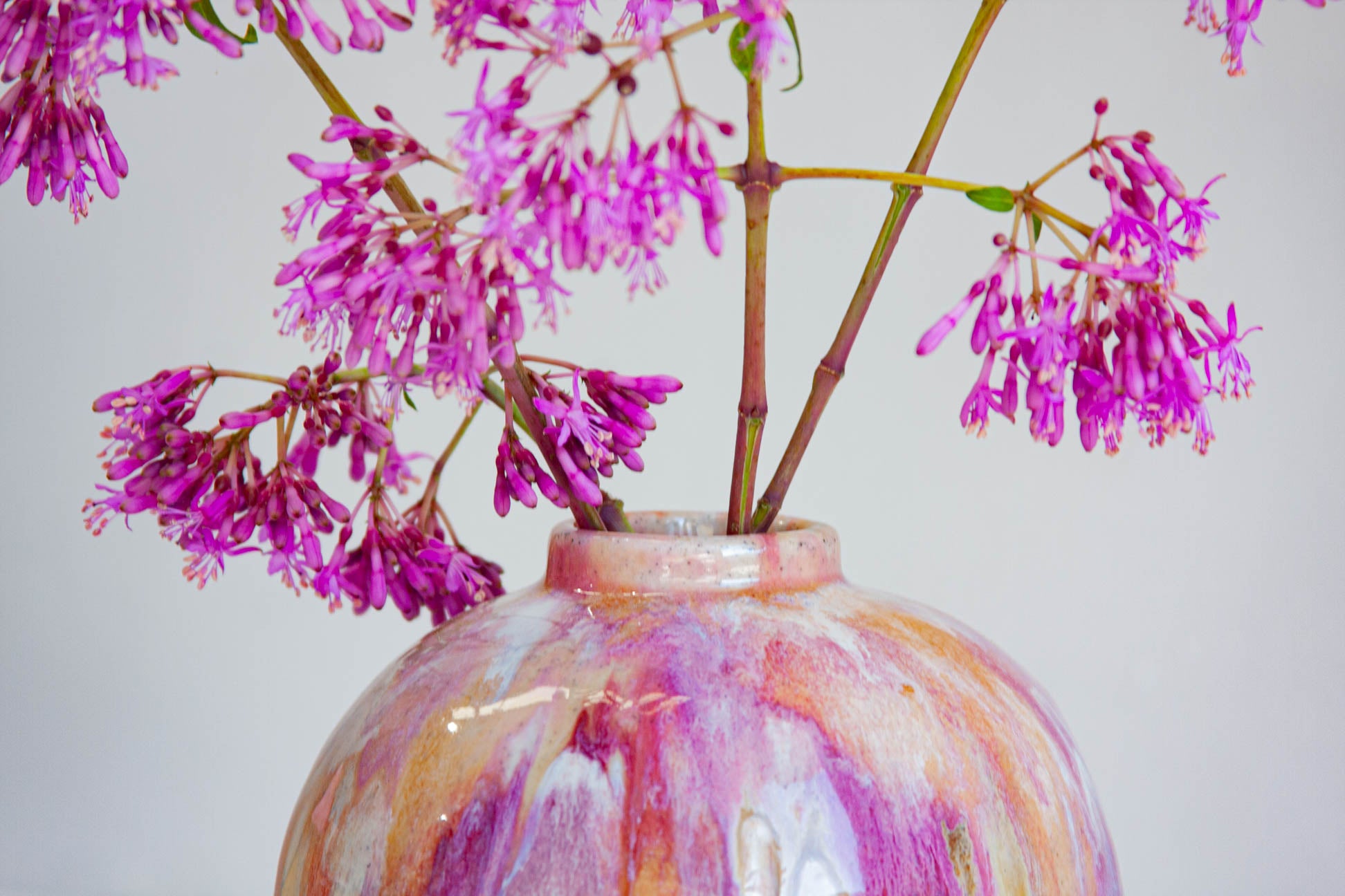 A close-up of a round ceramic vase with a marbled pink, purple, and cream glaze, holding pink-purple flowering branches against a plain light gray background.