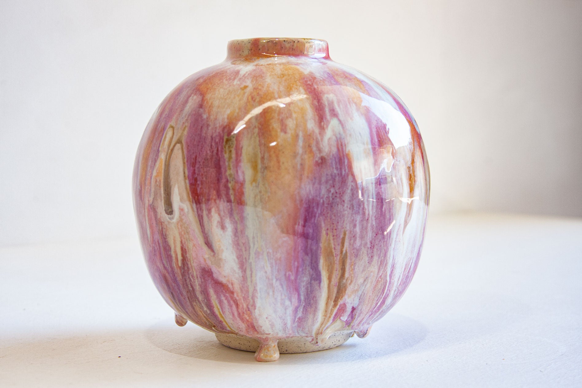 A small round ceramic vase with a glossy finish, featuring a marbled pattern of purple, orange, pink, and white glaze, standing on three small feet against a plain light background.
