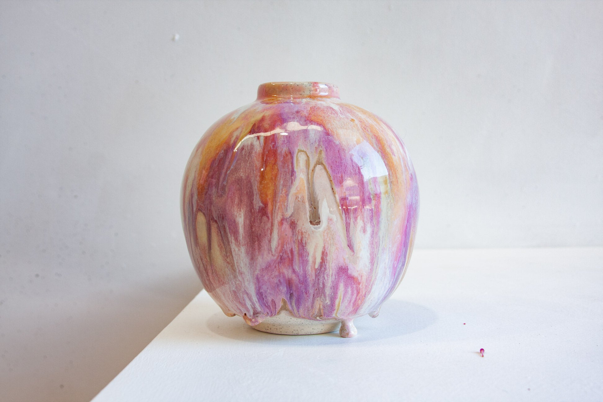 A small round ceramic vase with a glossy finish, featuring a marbled glaze of pink, purple, orange, and cream, displayed on a white surface against a light gray background.