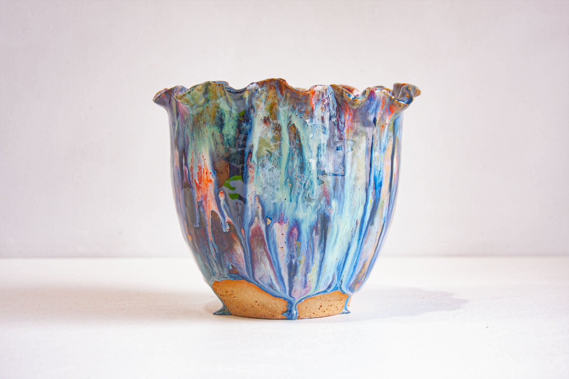 A handmade ceramic vessel with a ruffled rim, blue and teal glaze with orange and pink streaks running down to an unglazed speckled clay foot, on a white surface.