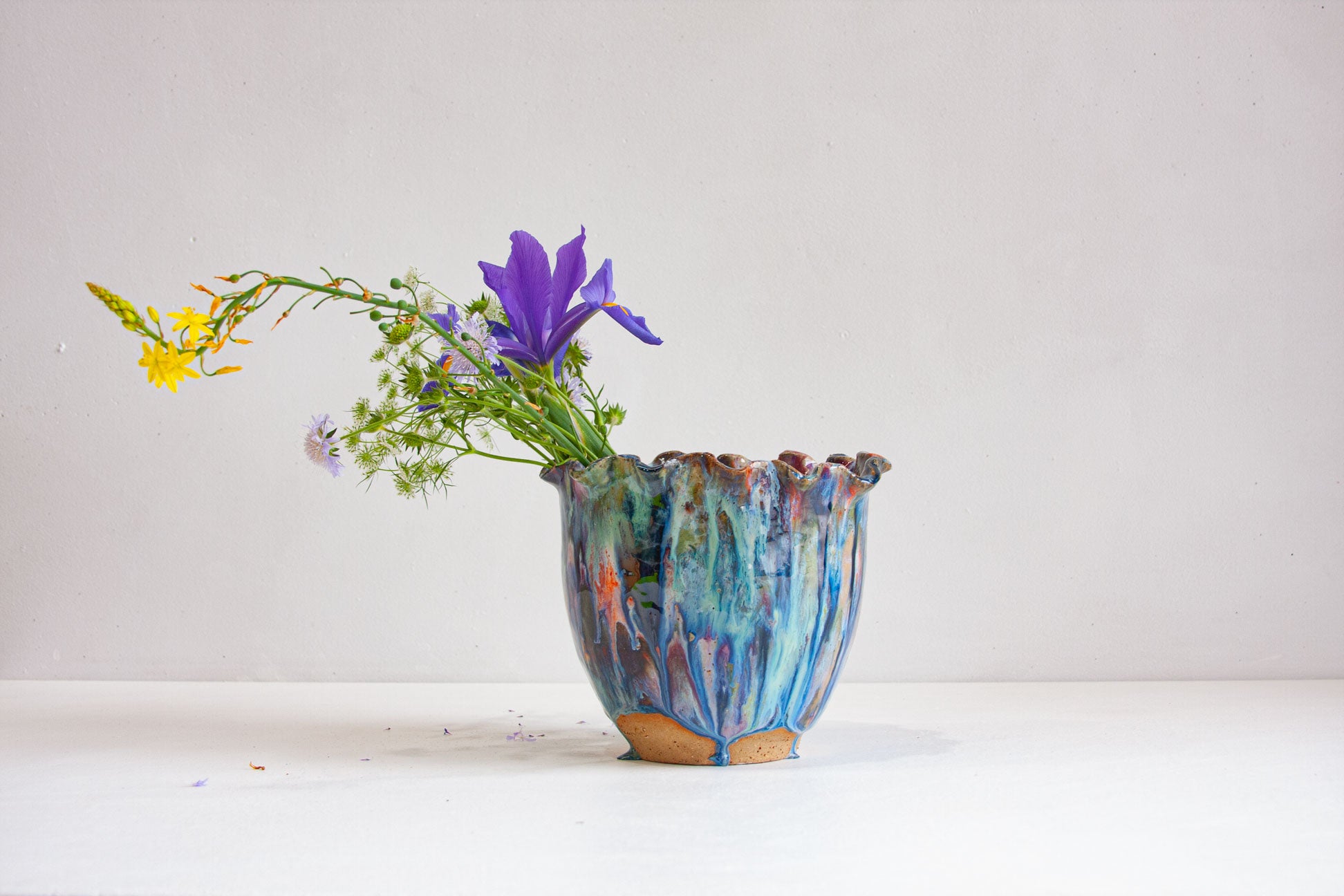 A handmade ceramic vessel with a ruffled rim, glazed in blue, teal, purple, pink and orange, holding purple iris, small yellow flowers and white scabiosa, on a white surface against a pale grey wall.
