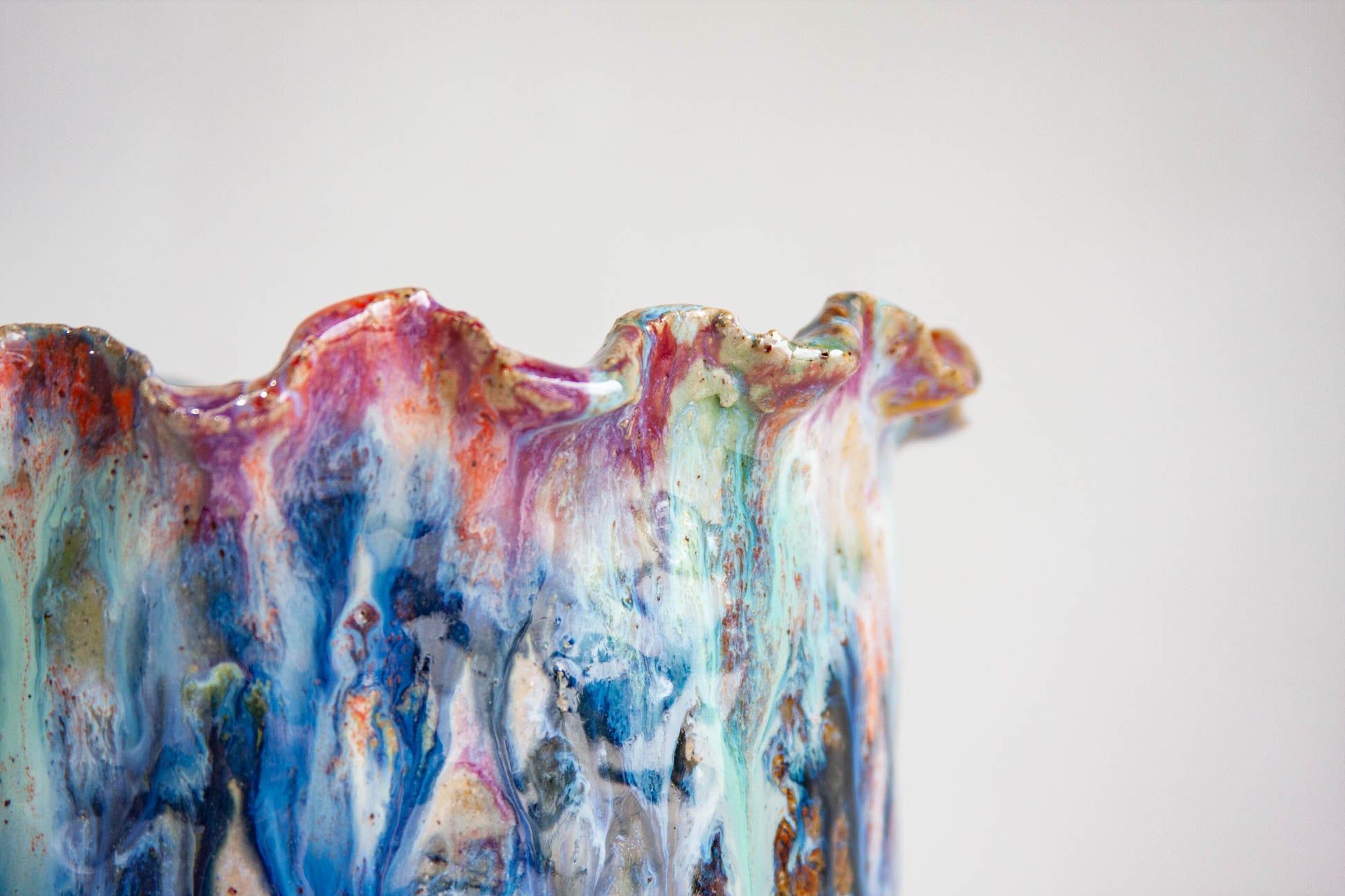 Close-up of the ruffled rim of a handmade ceramic vessel, with pink, purple, orange and warm brown gathering along the edge above vertical runs of blue, teal and cream.