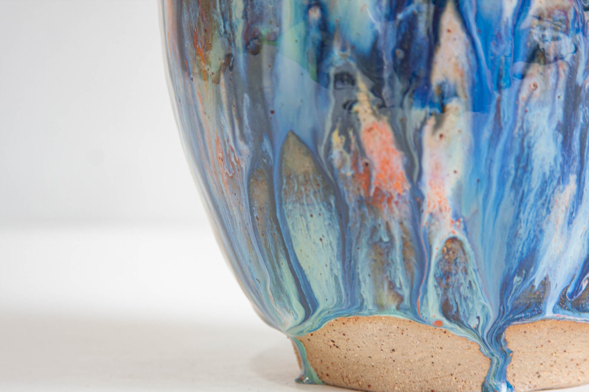 Close-up of the lower half of a handmade ceramic vessel, blue and teal glaze running in vertical streaks over coral orange, ending above a band of bare speckled clay at the foot.