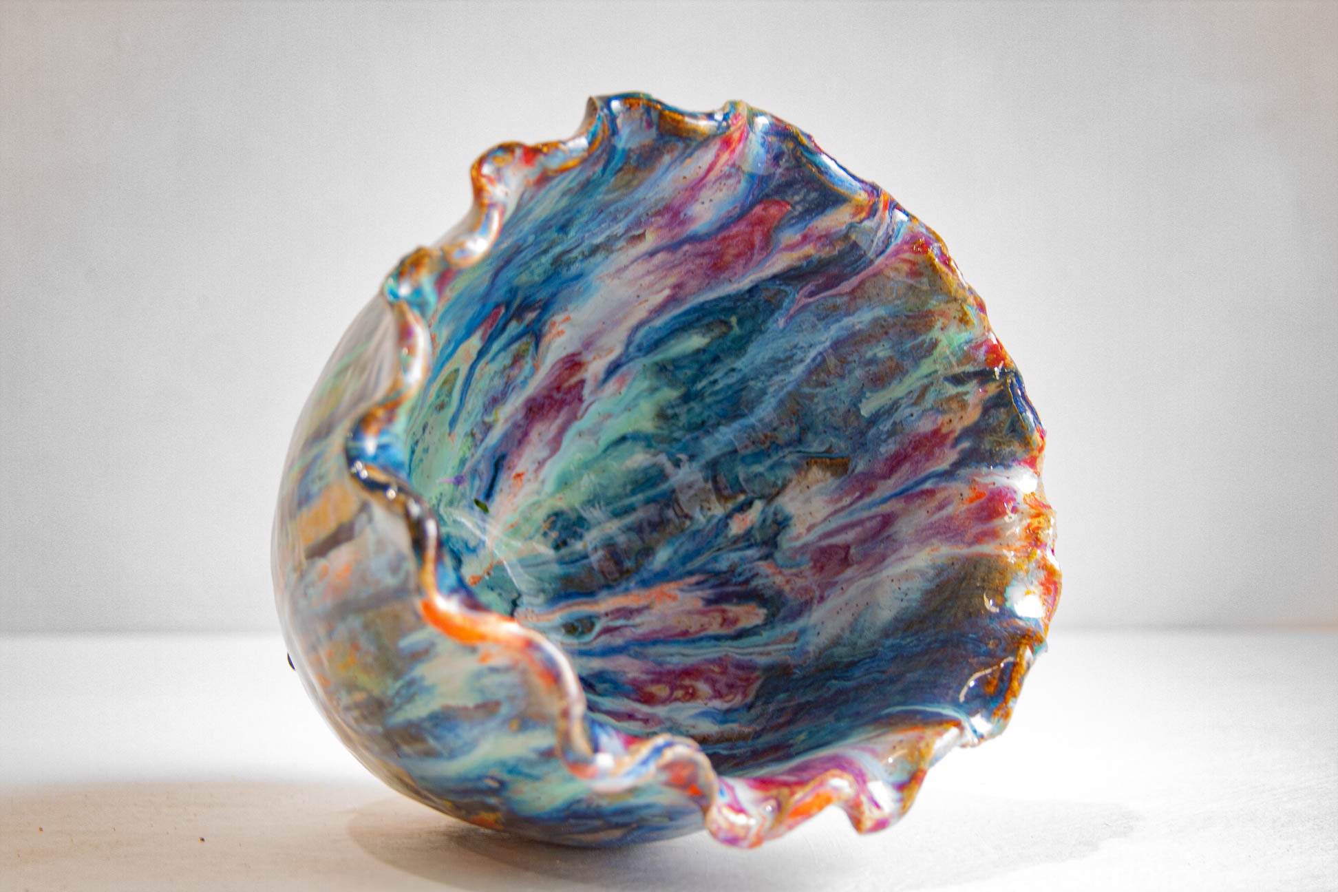 Looking down into a handmade ceramic vessel, showing the glazed interior in blue, teal, purple and pink, with the ruffled rim curving around it.