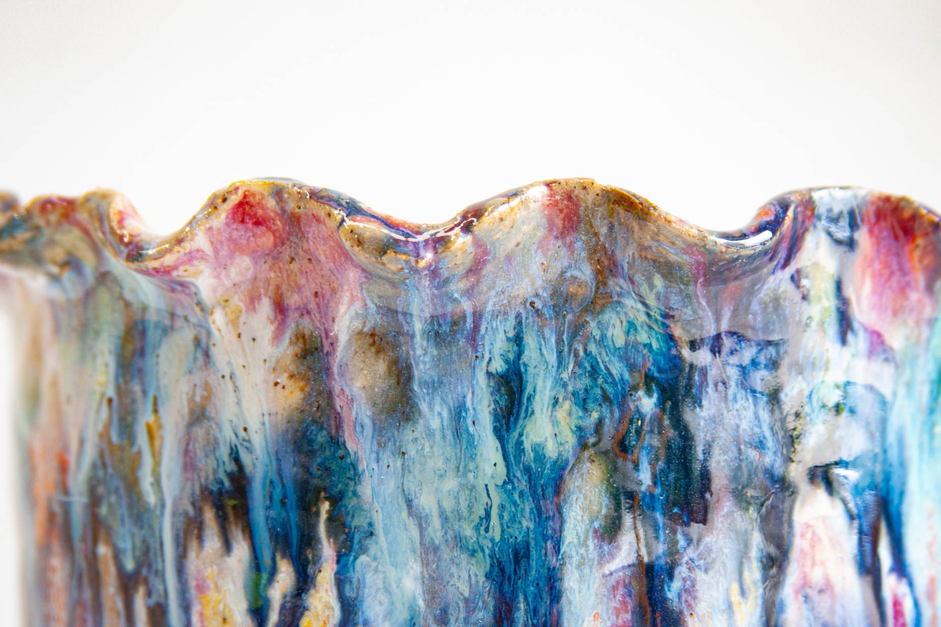 Close-up of the ruffled rim of a handmade ceramic vessel, warm brown and pink along the scalloped edge above streaks of blue, teal and white.