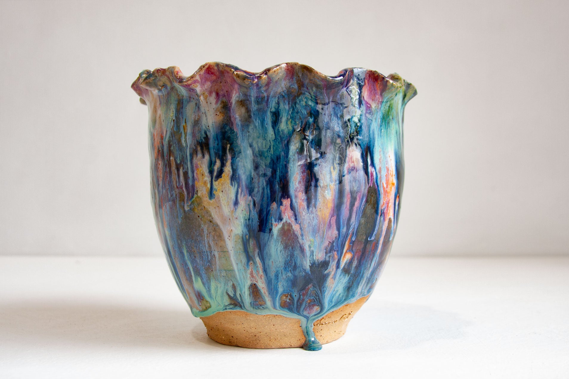A handmade ceramic vessel with a ruffled rim, glazed in blue, teal, purple, pink and orange, standing on a band of bare speckled clay, against a pale grey background.